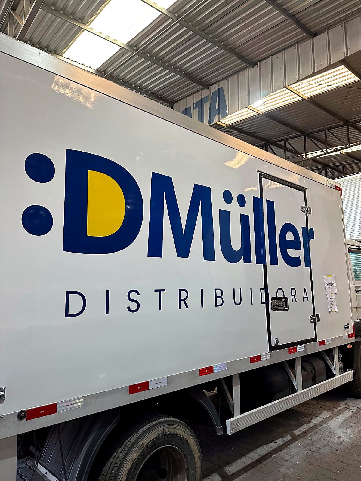 Caminhão baú branco com a logomarca D Müller Distribuidora em azul e amarelo