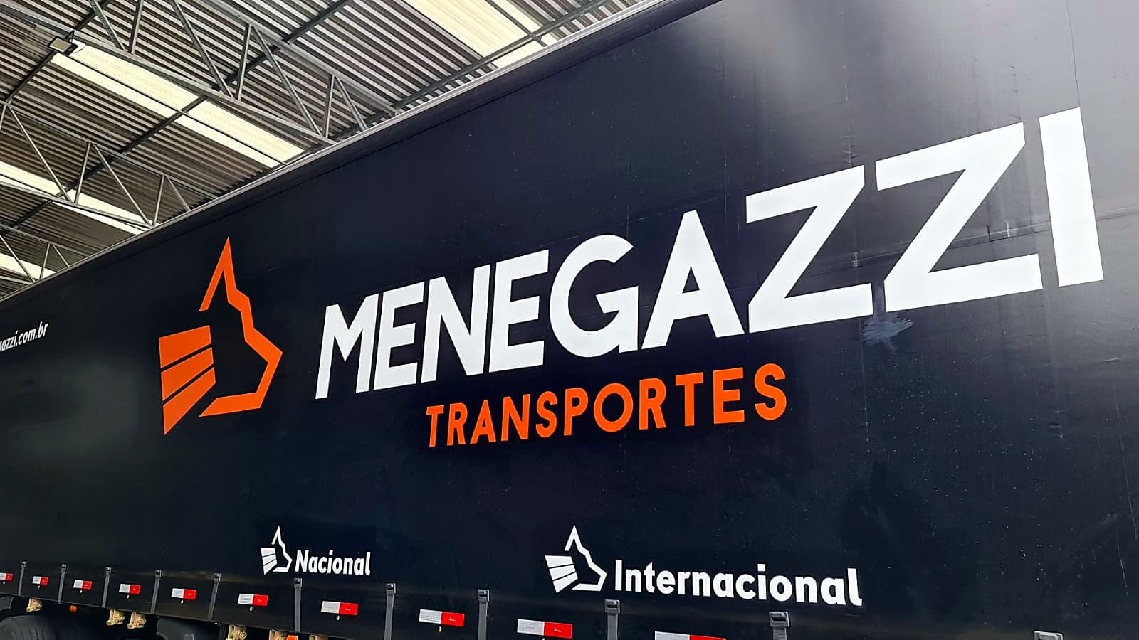 Lona preta de carreta sider com a logomarca Menegazzi Transportes em branco e laranja